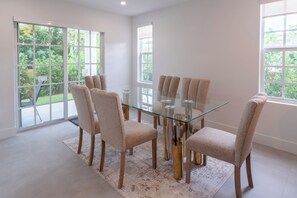 Un espace de salle à manger moderne avec une table en verre entourée de six chaises, de grandes fenêtres et une vue sur la verdure à l’extérieur.