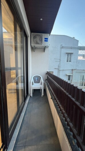 Een smal balkon met een witte plastic stoel, een raam met uitzicht op een gebouw en een airconditioning op de muur.