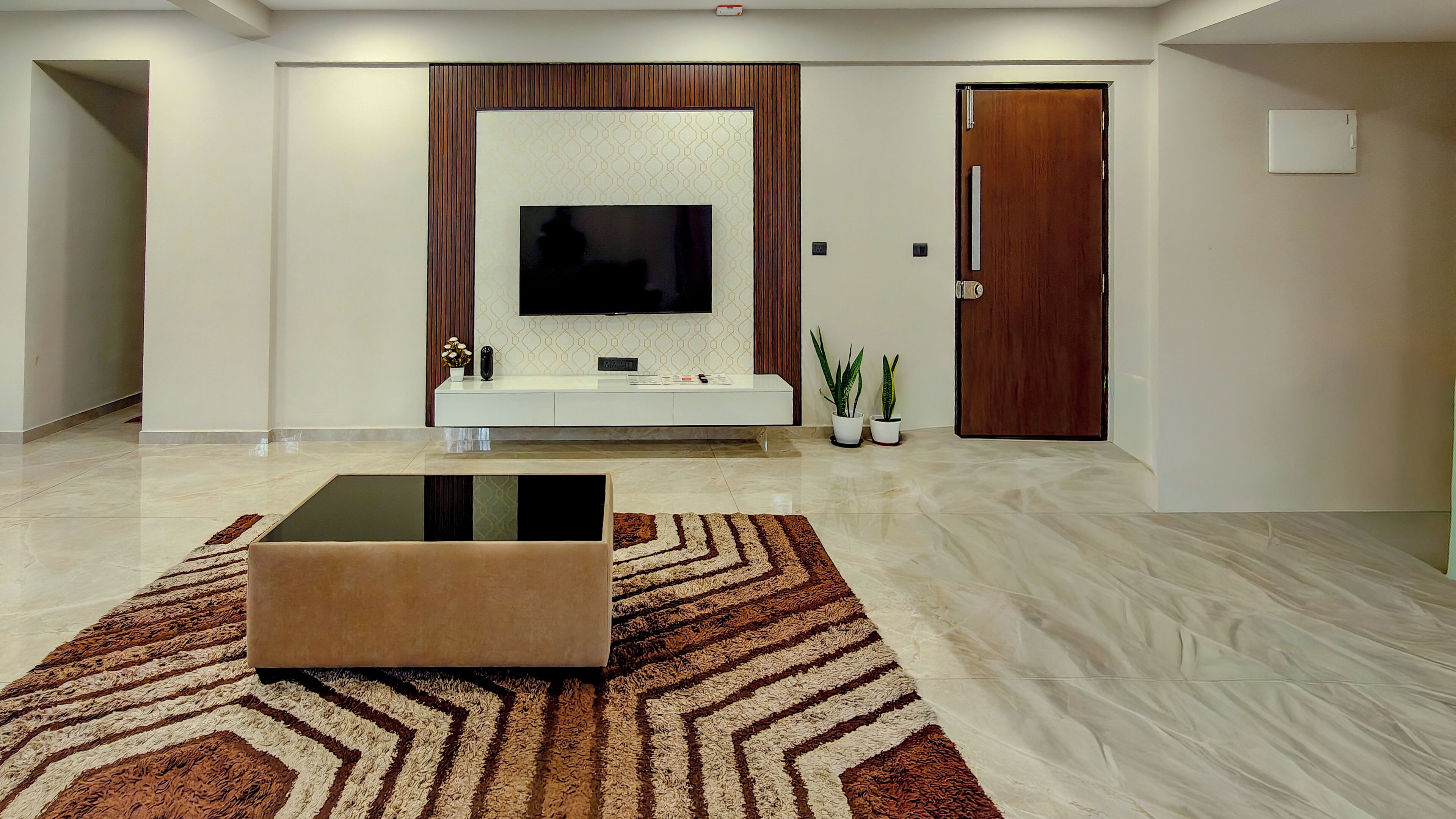 A modern living room with a flat-screen TV mounted on a wall, a white entertainment unit below, a brown sofa on a patterned rug, and a wooden door.