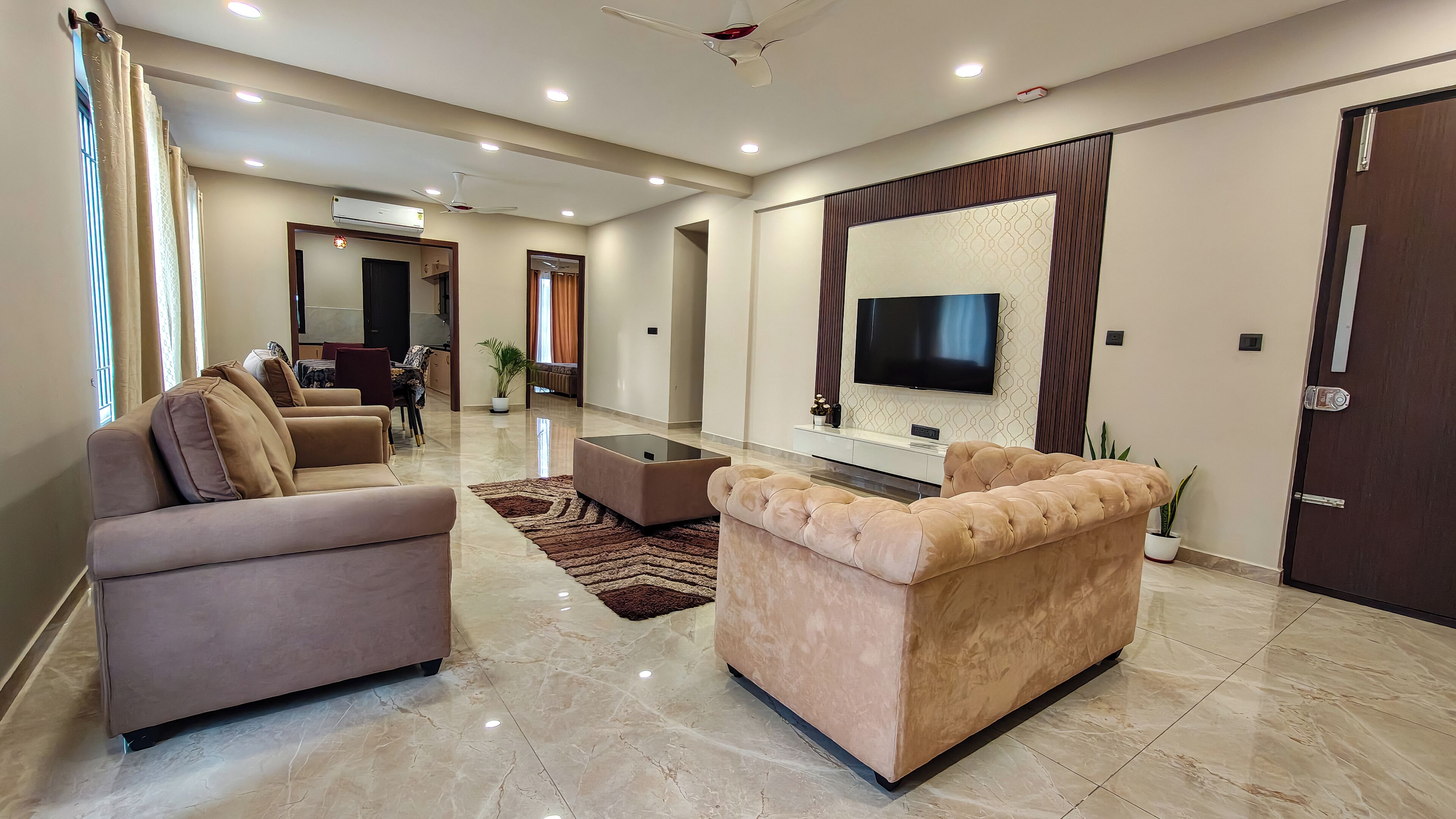 A spacious living room with a flat-screen TV mounted on the wall, a ceiling fan, and a large window with curtains.