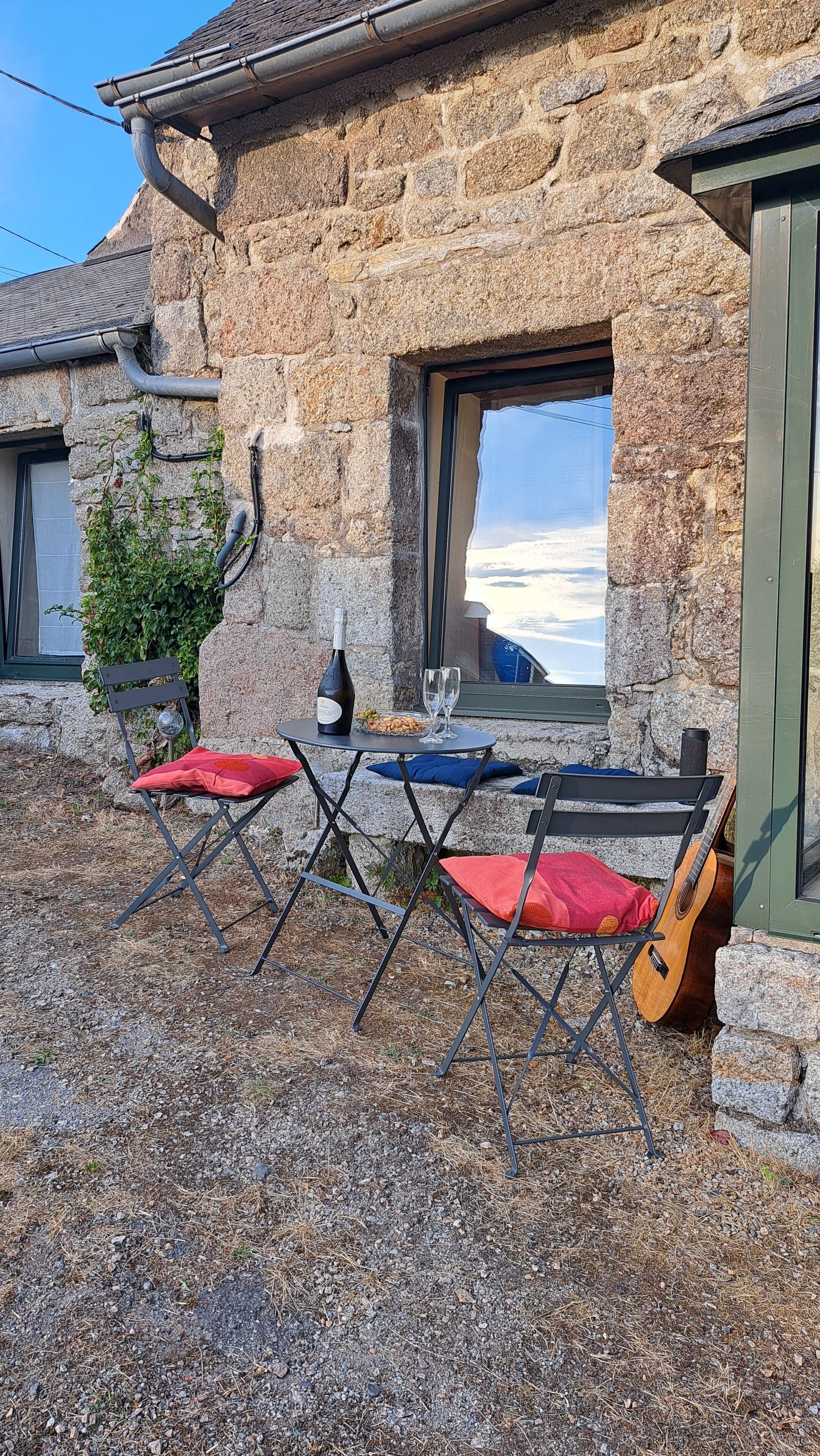 Ein Steingebäude mit einem Fenster, das auf eine verschneite Landschaft blickt, und ein kleiner Sitzbereich im Freien mit einem Tisch, zwei Stühlen, einer Gitarre und einer Weinflasche.