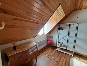 Eine schräge Decke mit Holzvertäfelung, ein Heizkörper, ein Stuhl mit Kissen, ein Regal mit Schuhen und ein kleiner Tisch mit einer Vase.
