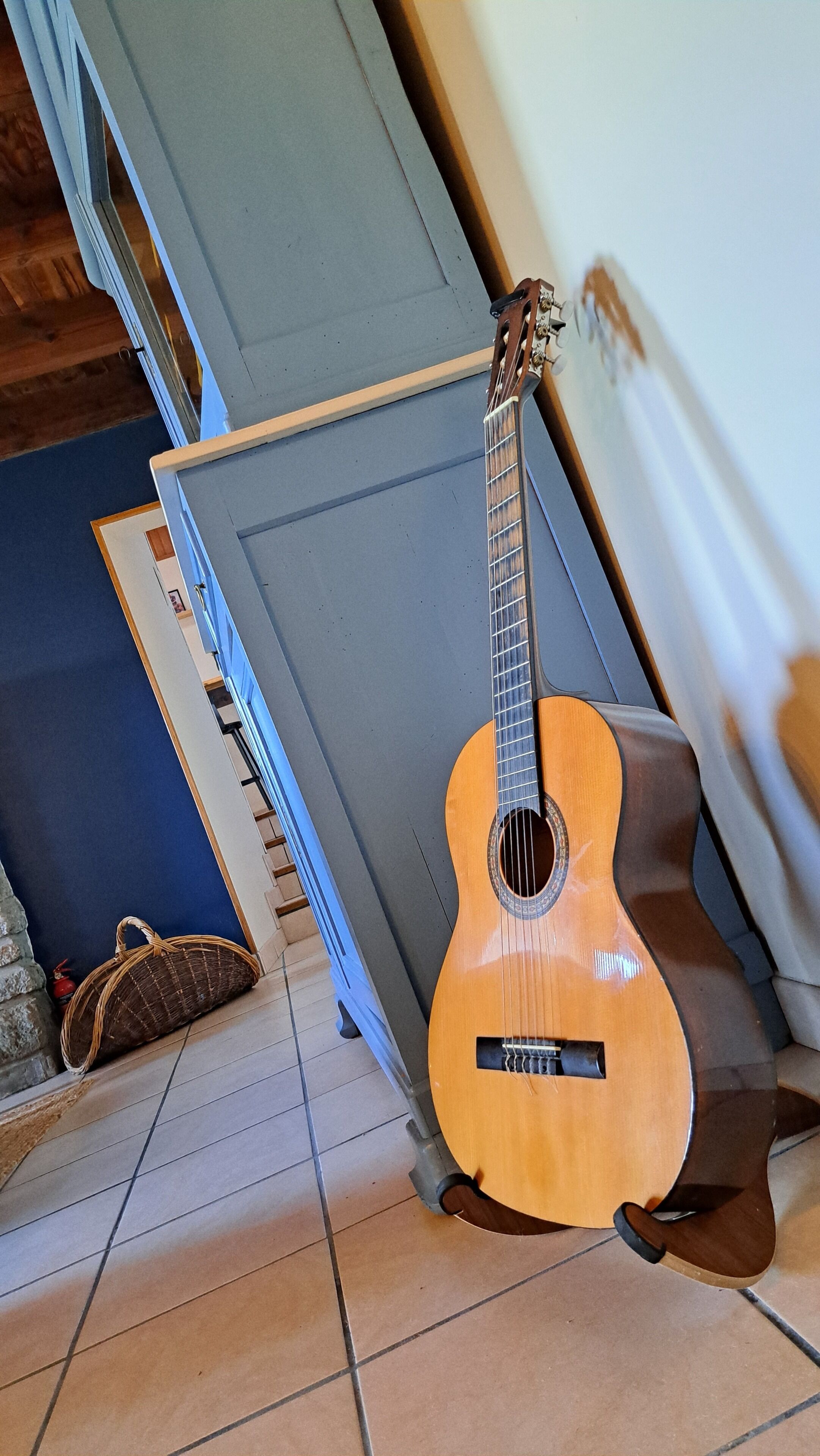 Eine Gitarre, die an einer Wand lehnt, ein blauer Stuhl und ein Korbwäscher auf einem Fliesenboden.