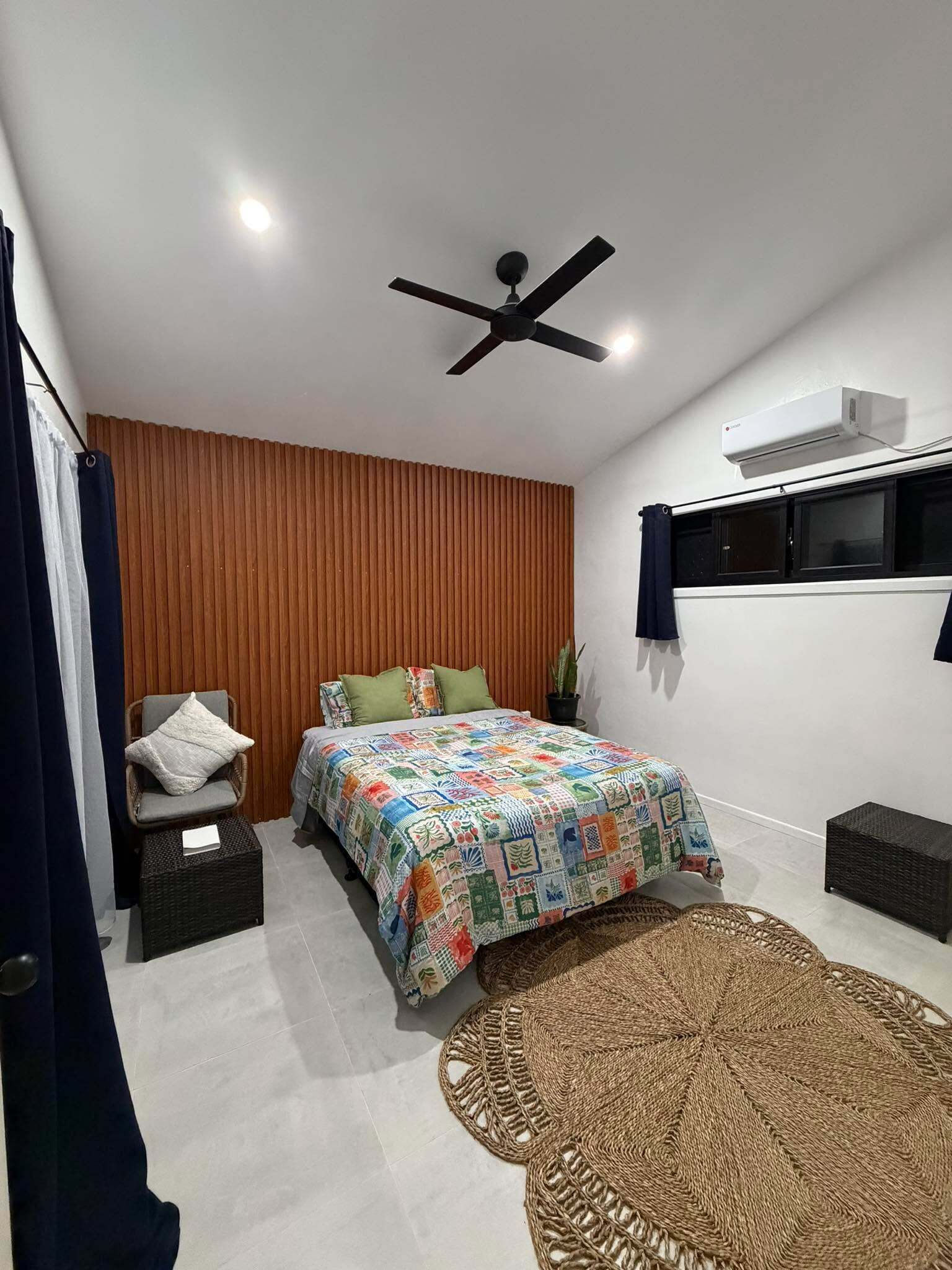 A bedroom with a bed, a ceiling fan, an air conditioning unit, a window with curtains, and a woven rug.