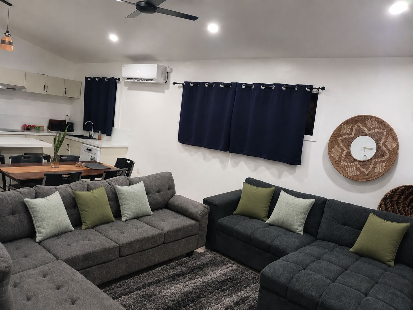 Een moderne woonkamer met grijze banken, een eettafel en een keuken met kasten en een airconditioning.