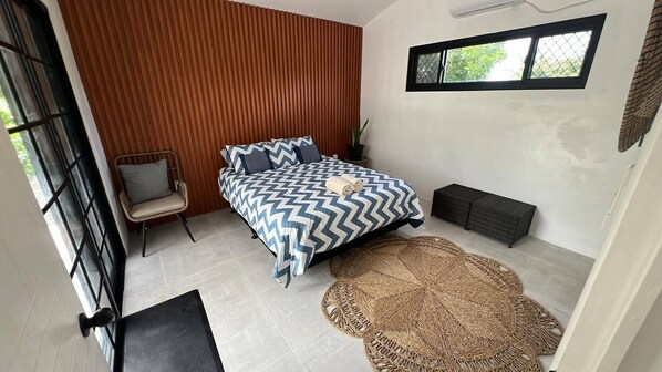 Un dormitorio con cama, una silla, un ventanal y un tapete tejido.
