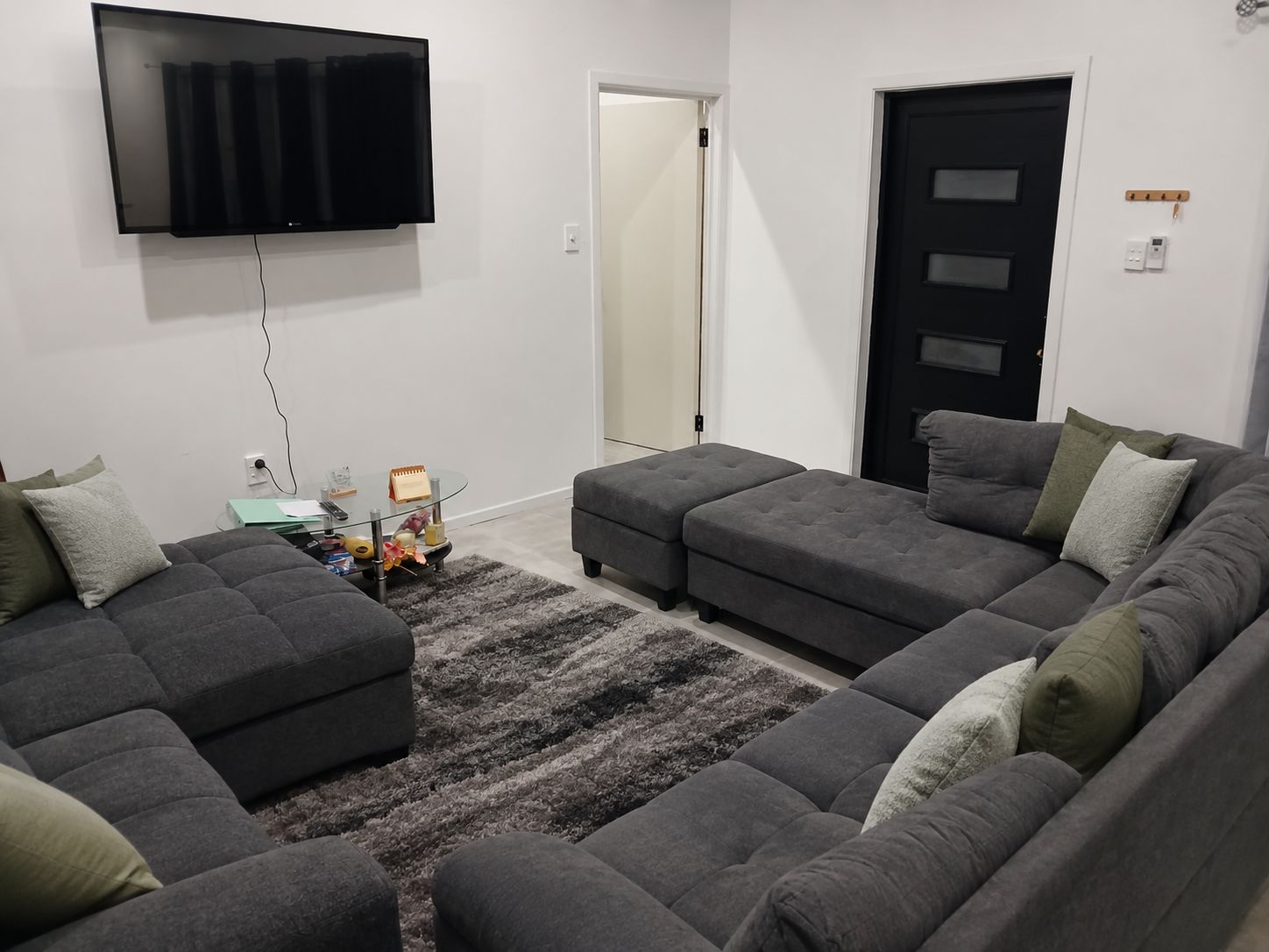 A modern living room with a sectional sofa, a flat-screen TV mounted on the wall, and a small side table with a glass top.
