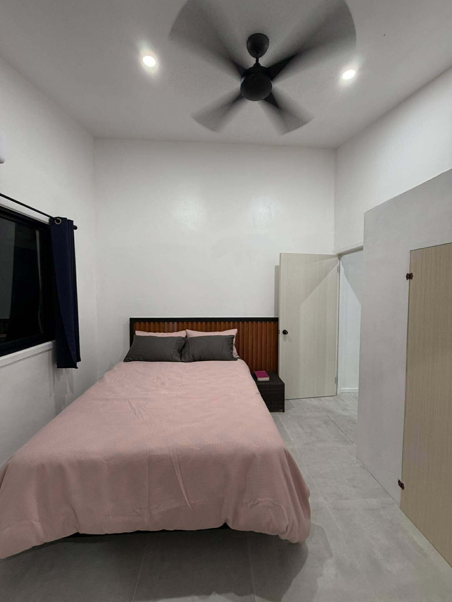 Um quarto com cama, ventilador de teto, janela com cortinas e porta.