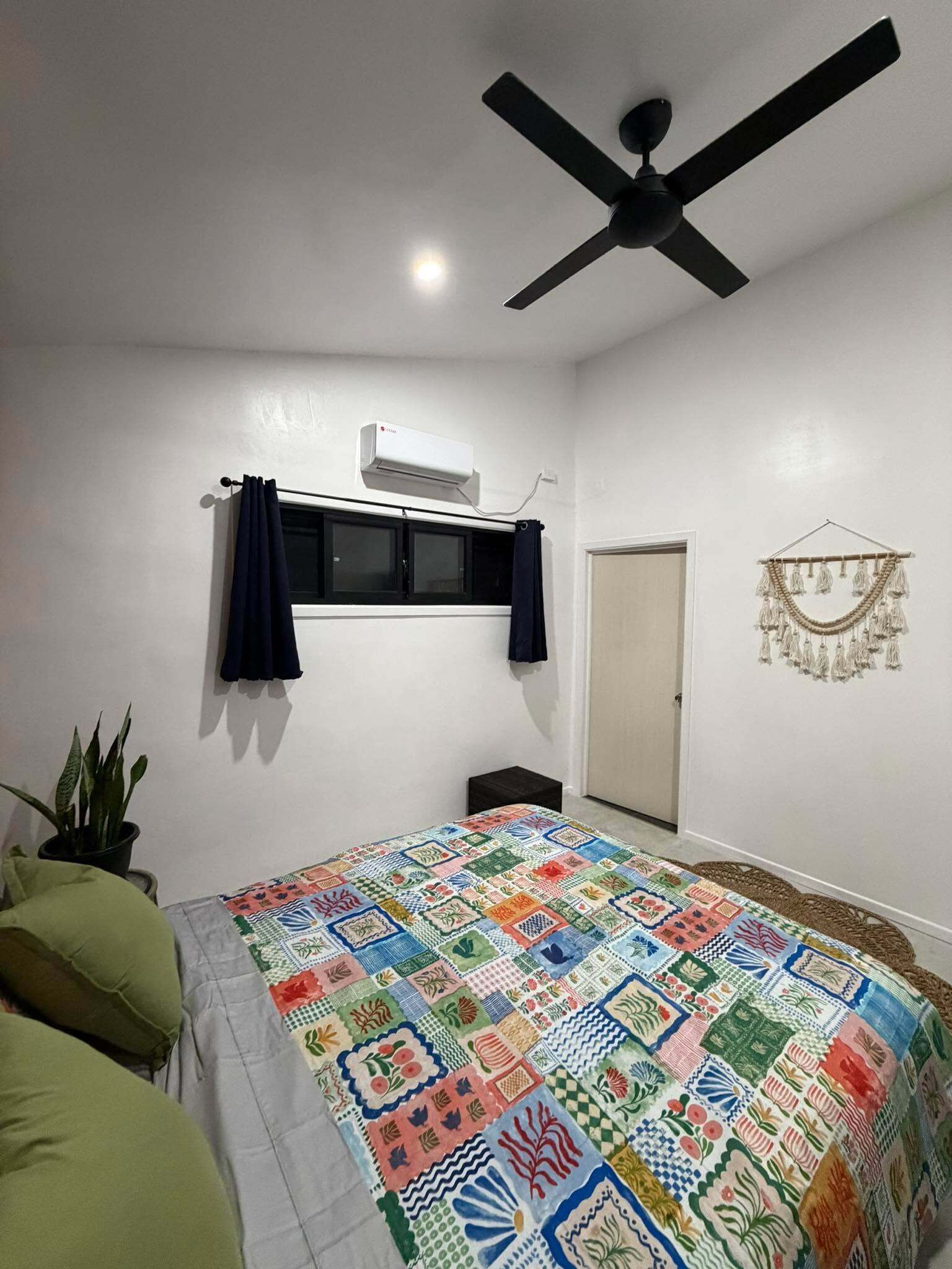 A bedroom with a bed, a ceiling fan, an air conditioning unit, a door, and a wall decoration.