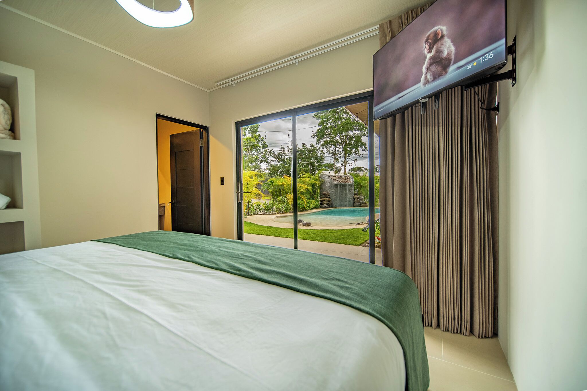 A hotel room with a bed, a television mounted on the wall, and a sliding glass door leading to an outdoor area with a pool and greenery.