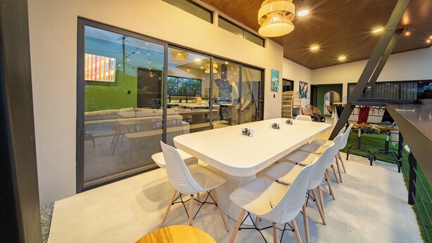 A modern dining area with a long table, white chairs, and a view through large glass doors. - Casa Nantú - Arenal Volcano Views • Zero-Entry Pool • Sauna • 4BR/4.5BA (La Fortuna)