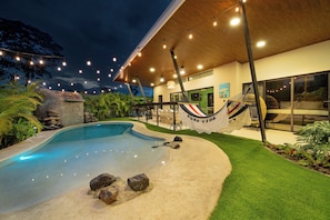 A modern outdoor pool area with a hammock, a dining area, and a view of the house. - Casa Nantú - Arenal Volcano Views • Zero-Entry Pool • Sauna • 4BR/4.5BA (La Fortuna)