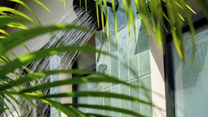 A modern building with a rain shower head, a window with a view, and a palm plant in the foreground. - Casa Nantú - Arenal Volcano Views • Zero-Entry Pool • Sauna • 4BR/4.5BA (La Fortuna)