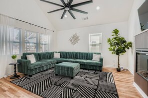 A modern living room with a sectional sofa, a ceiling fan, a large window with blinds, and a potted plant. - 5 king beds, 2 kitchens, 2 living rooms | East Atlanta Village (Atlanta)
