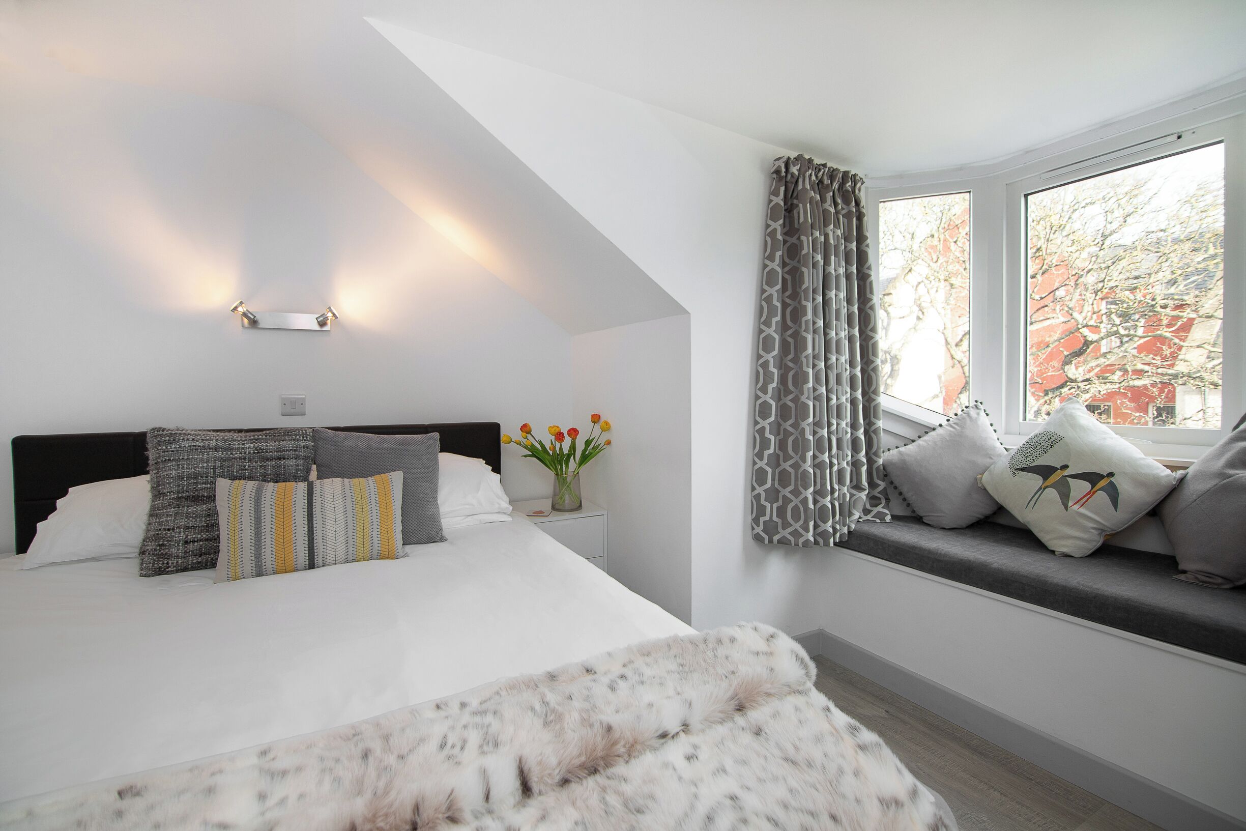 A modern bedroom with a large bed, a bedside table with a vase of flowers, and a window with patterned curtains.