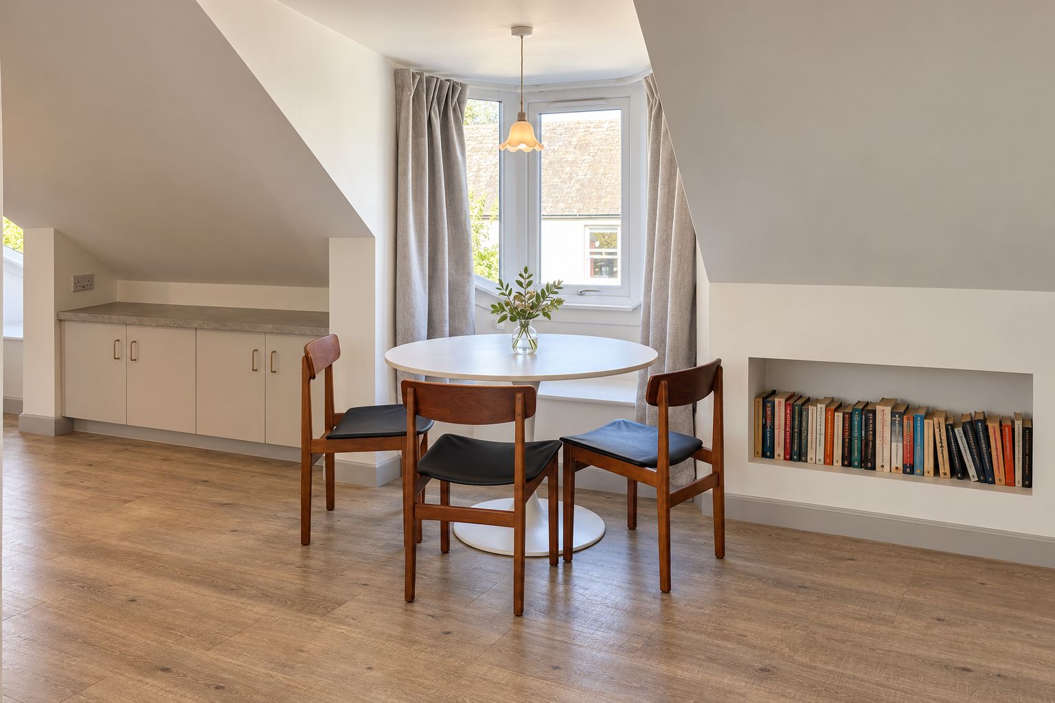 A dining area with a round table, four chairs, a vase with greenery, and a bookshelf with books.