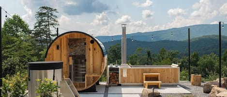 A wooden sauna structure with a stone staircase, a bench, and a small table, set against a mountainous backdrop.