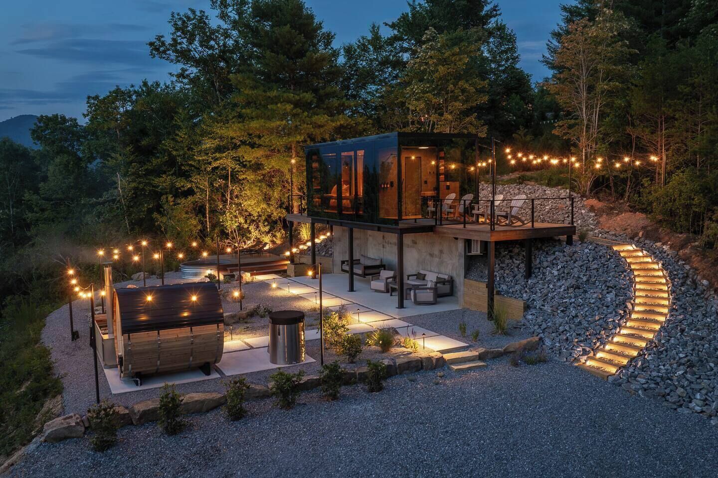 Un espacio exterior moderno con terraza, zona de estar y una pequeña construcción con paredes de vidrio.