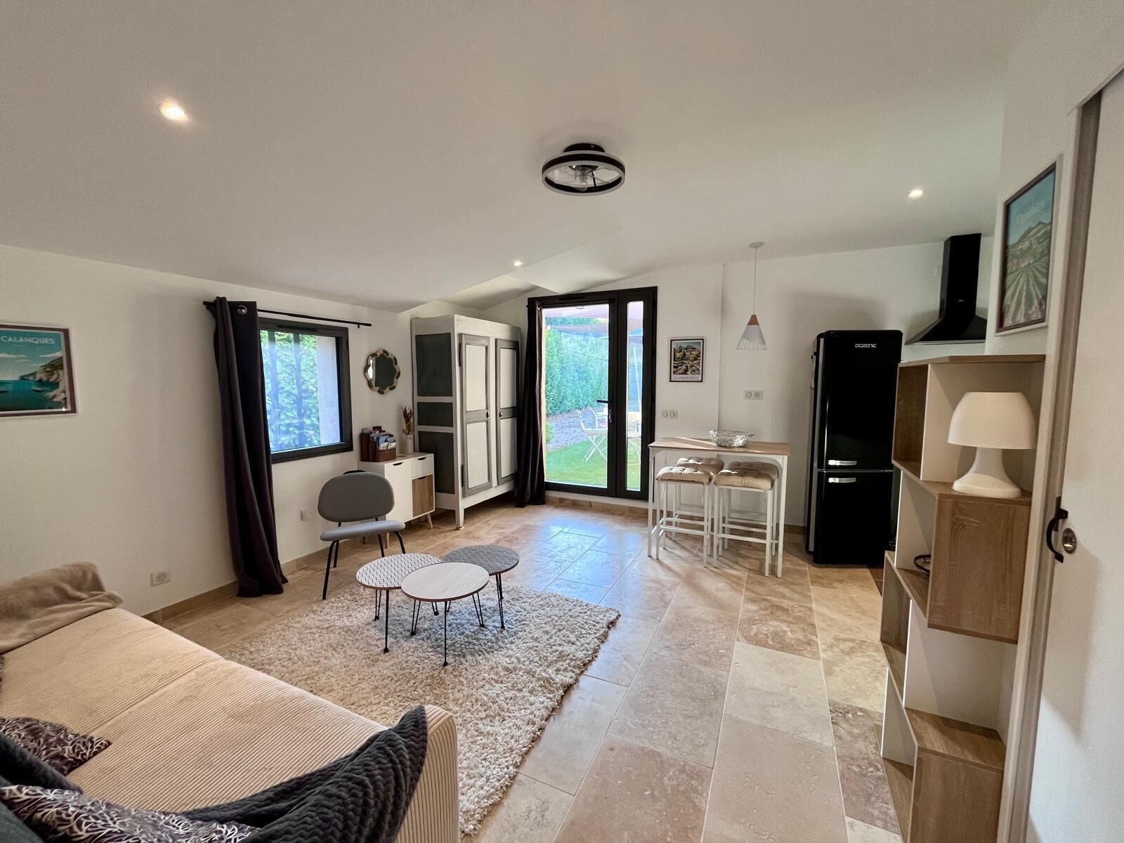 A spacious living area with a sofa, a dining table with chairs, a kitchen area with a refrigerator, and a sliding glass door leading to a garden.