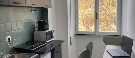 Een kleine keuken met een eethoek, een raam met uitzicht op bomen, en een laptop op tafel.