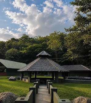 A pavilion with a tiled roof, surrounded by a grassy area and trees. - Hills and Chills Nature Stay (Sakleshpur)