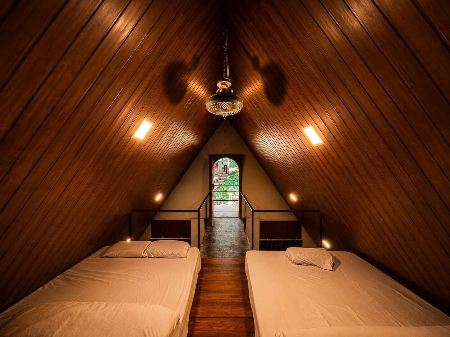 A triangular attic bedroom with two beds, wooden slats on the ceiling, and a staircase leading to a balcony.