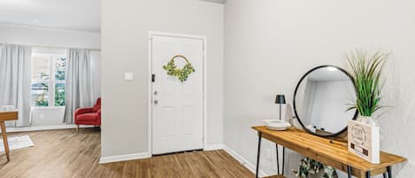 A hallway with a wooden floor, a white door with a green wreath, a mirror on a wooden console table, and a red chair.