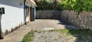A courtyard with a stone wall, a paved area, and a building with a door and shutters.