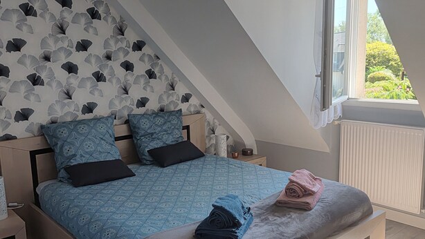 Ein Schlafzimmer mit schrägen Wänden, einem Bett mit blau gemustertem Bettbezug, zwei Kissen und einem Nachttisch mit Lampe.