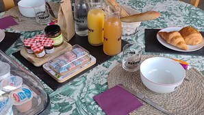 Ein Esstisch mit floralem Tischläufer, ein Holzstuhl, ein Croissant, ein Glas Orangensaft, eine Saftflasche, ein Marmeladenglas und eine Schüssel.