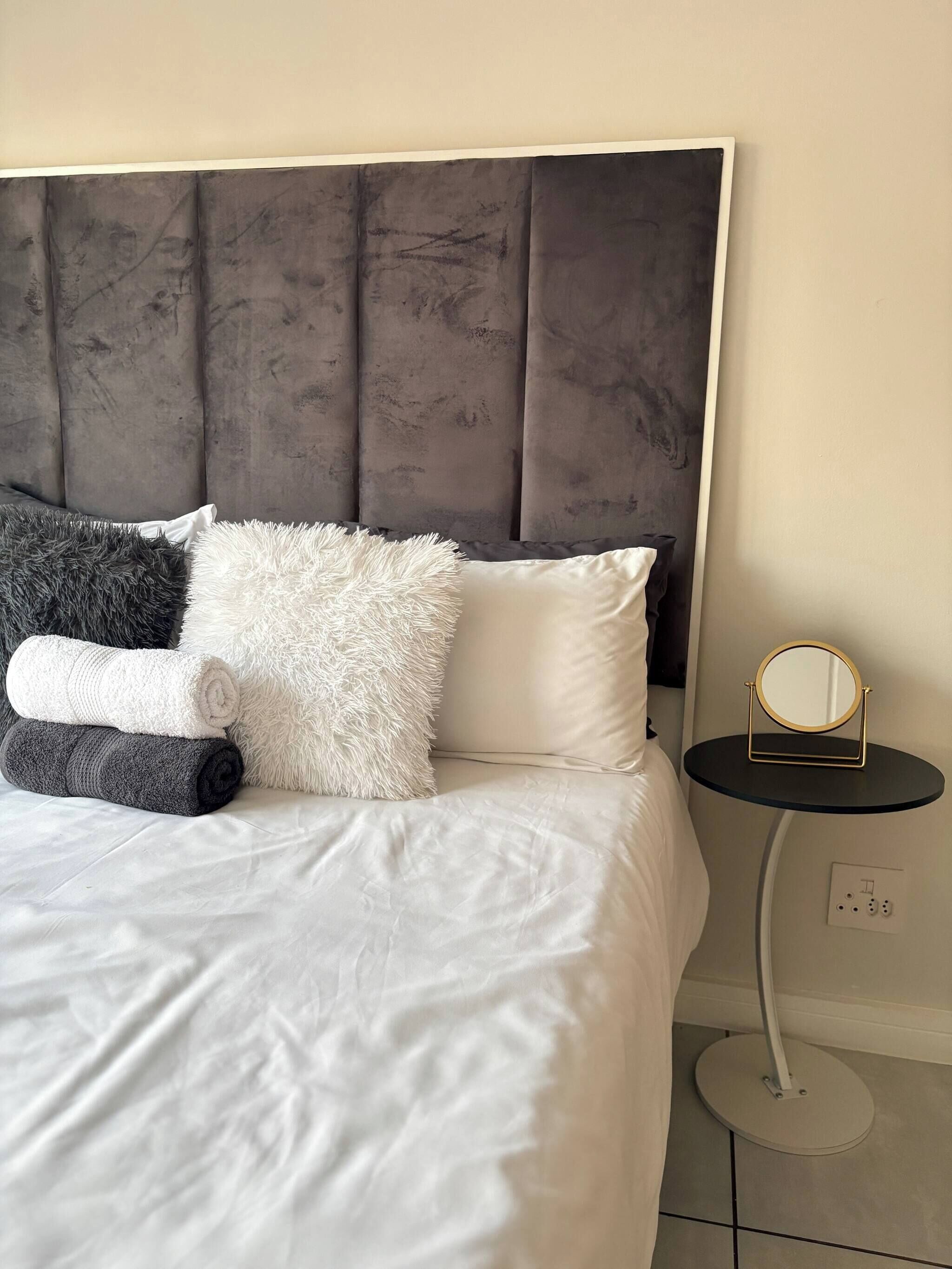 Ein ordentlich bezogenes Bett mit grauem Polsterkopfteil, einem weißen und grauen flauschigen Kopfkissen und einem weißen Kissen. Ein kleiner runder Tisch mit goldfarbener Spiegelrahmung steht neben dem Bett.