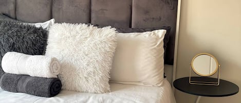 Una cama bien hecha con un cabecero tapizado gris, un cojín blanco y gris esponjoso, y un cojín blanco. Una mesita redonda con espejo de marco dorado está al lado de la cama.