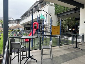 An outdoor seating area with tables and chairs, a playground with a slide, and a building with a balcony in the background. - Luxurious 2-bedroom Apartment in Pretoria With Pool Access (Pretoria)