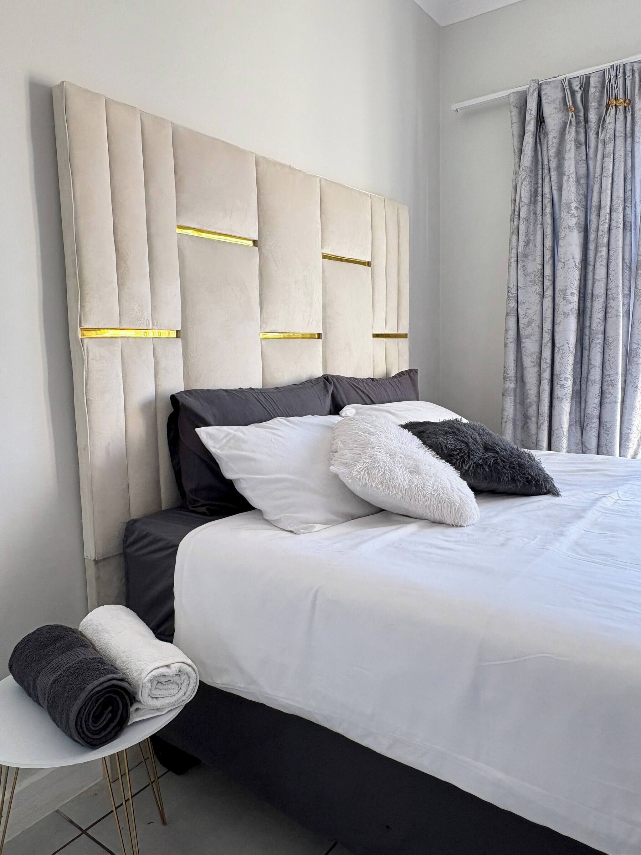 A modern bedroom with a tufted headboard, a bed with white linens, and a side table with rolled towels.
