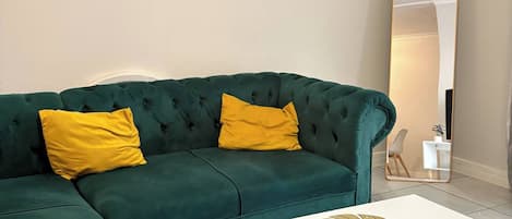 A green sectional sofa with yellow cushions, a white coffee table with a floral design, and a large mirror on the wall.