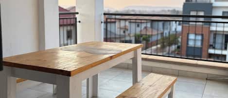 A balcony with a wooden table and bench, a tiled floor, and a view of buildings in the distance.