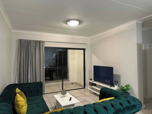 A modern living room with a sofa, a coffee table, a flat-screen TV, and a balcony with a railing. - Luxurious 2-bedroom Apartment in Pretoria With Pool Access (Pretoria)