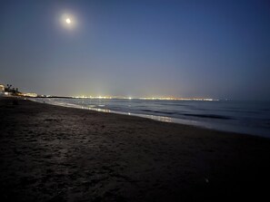 Een strand 's nachts met een brug in de verte en lichtjes van de stad langs de kustlijn.