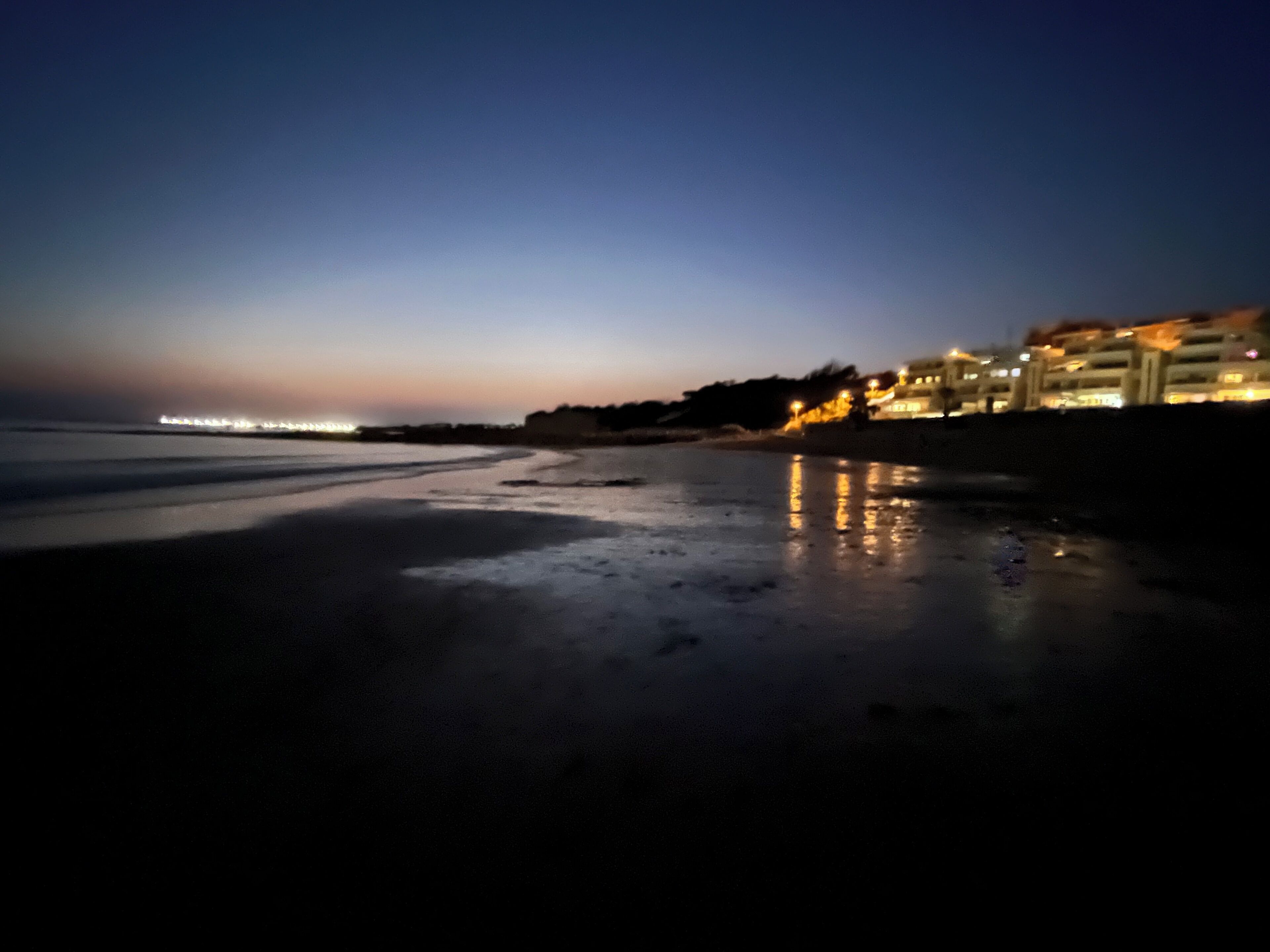 A beach at dusk with wet sand and a distant cityscape.