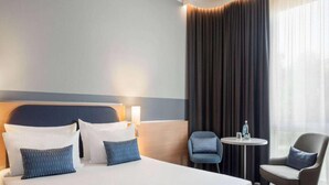 Una habitación de hotel con una cama grande, dos sillas, una mesita y un ventanal con cortinas.