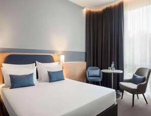 Quarto de hotel com uma cama grande, duas cadeiras, uma mesa pequena e uma janela com cortinas.