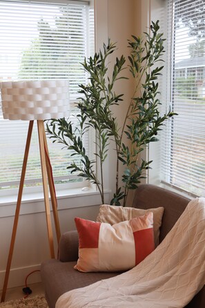 A room with a window, a plant, a chair with cushions, and a floor lamp.