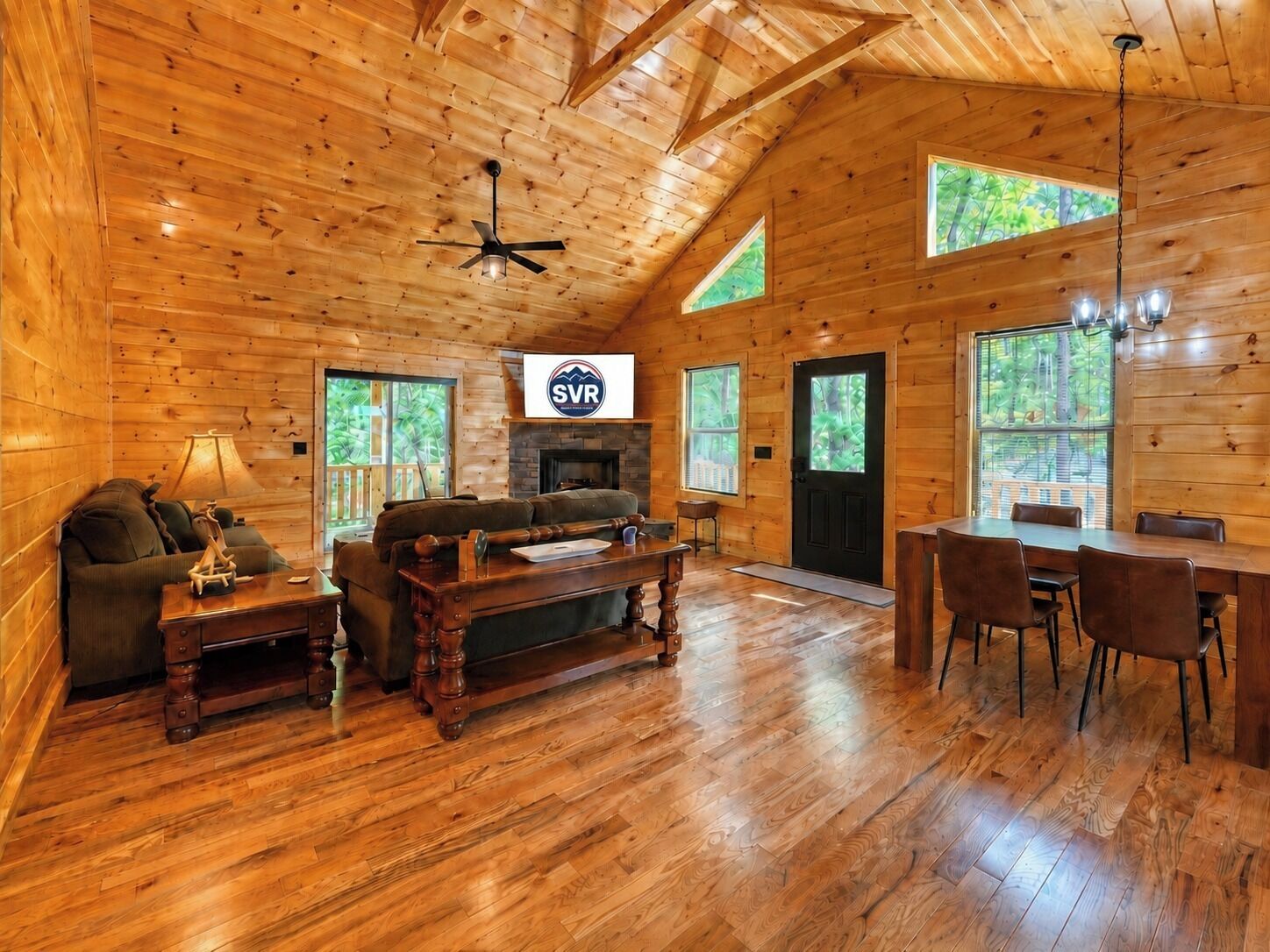 A log cabin interior with a fireplace, dining area, and a ceiling fan.