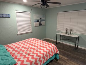 A bedroom with a bed, a ceiling fan, two windows with blinds, a small table with decorative items, and a painting on the wall. - Papa Ric's Happy Place (Bolivar Peninsula)