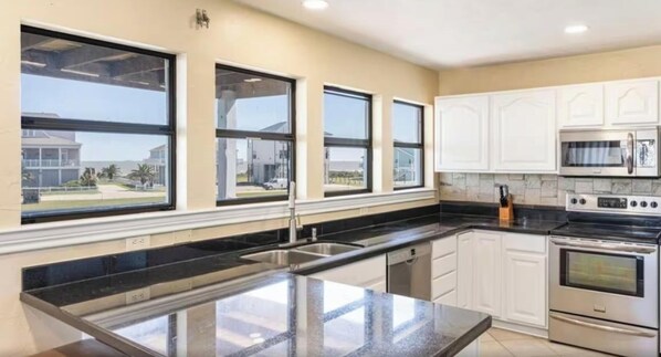 A kitchen with a large island, stainless steel appliances, and a view of a residential area. - Papa Ric's Happy Place (Bolivar Peninsula)