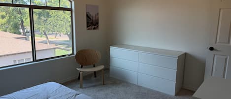 A bedroom with a bed, a chair, a dresser, and a window with a view of trees and a building.