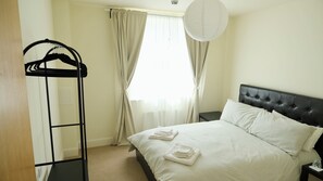 A neatly made bed with folded towels, a black hanging rack, and a window with curtains. - Quiet refurbished flat by Fulham Broadway (London)