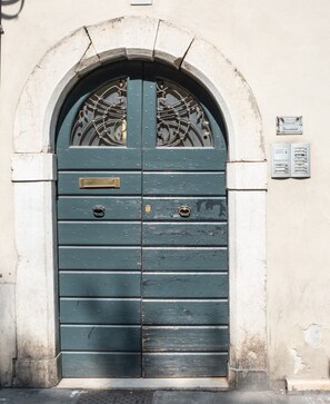 A green double door with a metal grille, set in a stone archway, with a utility box to the right. - [Historic Center] Elegant one-bedroom apartment with a balcony (Brescia)
