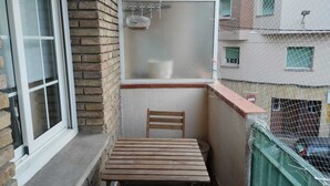 A small balcony with a wooden table and chair, a window with a screen, and a view of a multi-story building. - Barcelona Room – Temporary Sublet €40/day: 10% discount for longer stays (Spain)
