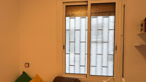 A bedroom with a bed, pillows, a bedside table with a lamp, and a window with a view of a brick wall. - Barcelona Room – Temporary Sublet €40/day: 10% discount for longer stays (Spain)